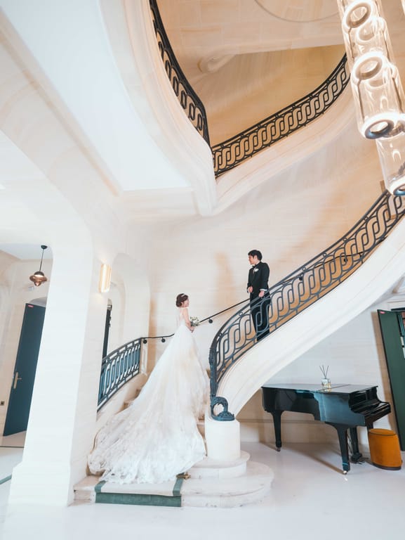 The bride climbs the curved stone staircase of a French château towards the groom, grand piano below