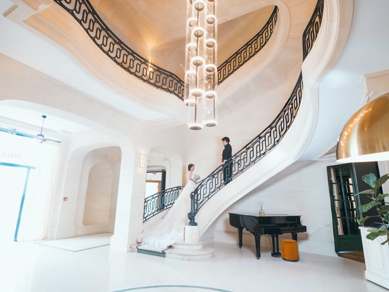 Le marié en haut du grand escalier à rampe de fer forgé du Château Mont Royal, la mariée au pied des marches près du piano