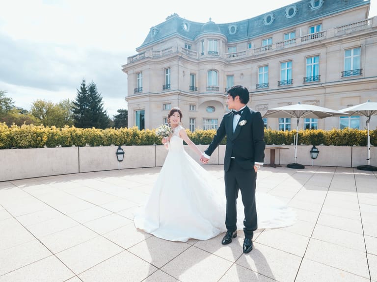 Les mariés main dans la main sur la terrasse devant la façade du Château Mont Royal, ciel bleu et haies taillées