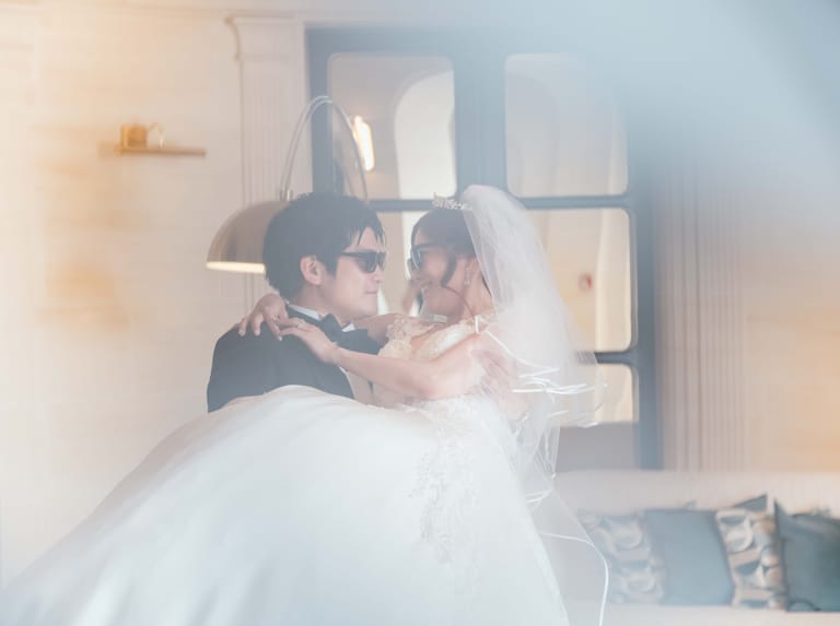 The groom in sunglasses lifts the veiled bride in a château lounge, shot through a blurred railing