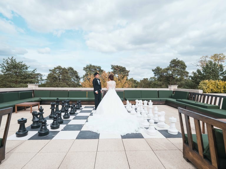 La mariée au milieu de l'échiquier géant du toit-terrasse, le marié face à elle, pins et ciel nuageux