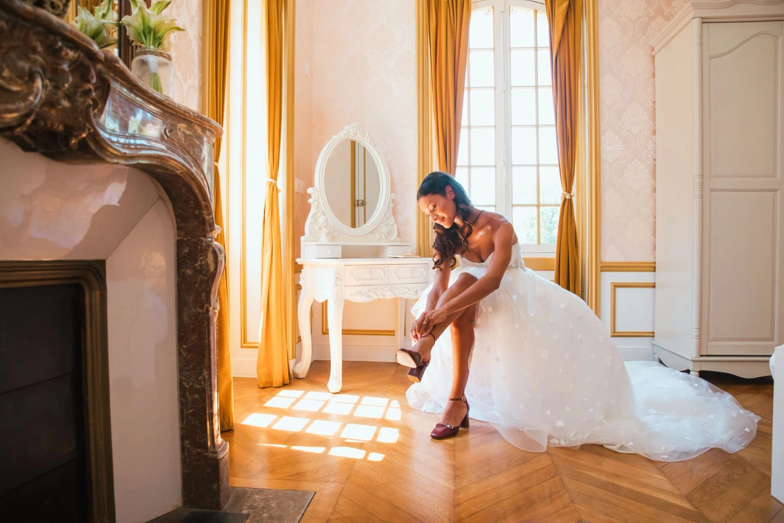 La mariée assise devant la coiffeuse à miroir ovale enfile ses escarpins bordeaux, lumière de fenêtre sur le parquet à chevrons, mariage au Château de Moliens, par Jean-Baptiste Argyre (Jaysphotography)