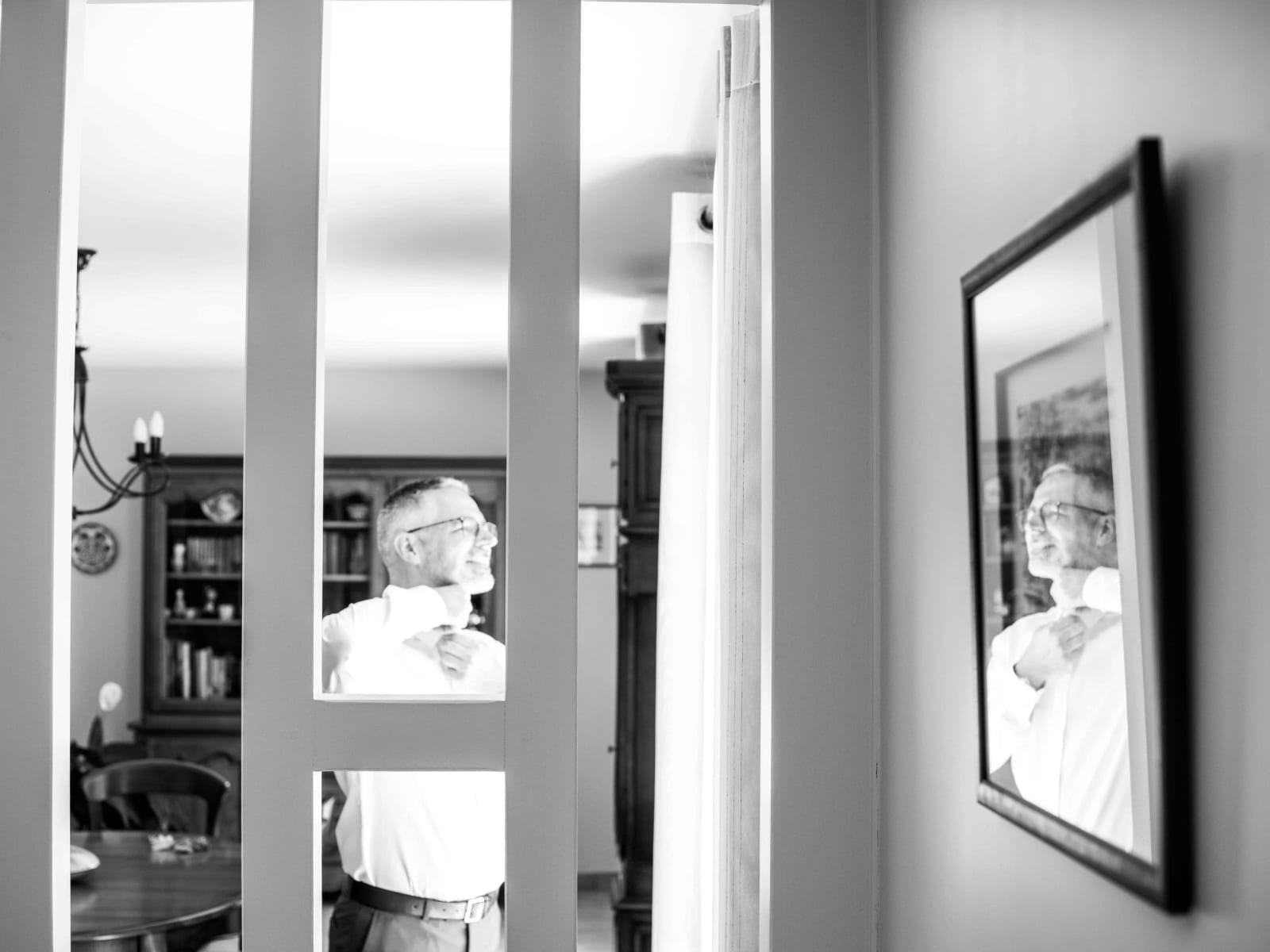 Éric ajuste le col de sa chemise devant le miroir pendant ses préparatifs, en noir et blanc, mariage au Domaine la Ferme du Roy, par Jean-Baptiste Argyre (Jaysphotography)