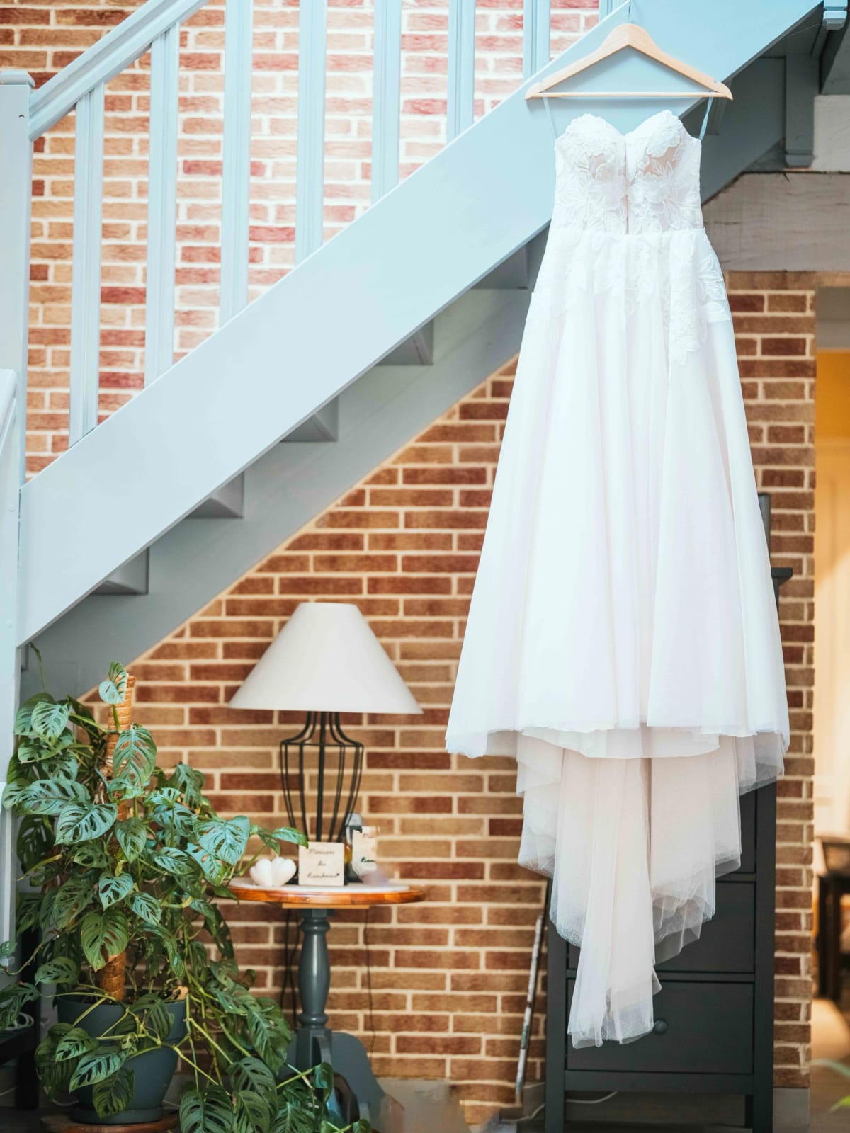 La robe de mariée en dentelle suspendue à la rampe de l'escalier devant un mur en briques, mariage au Domaine la Ferme du Roy, par Jean-Baptiste Argyre (Jaysphotography)
