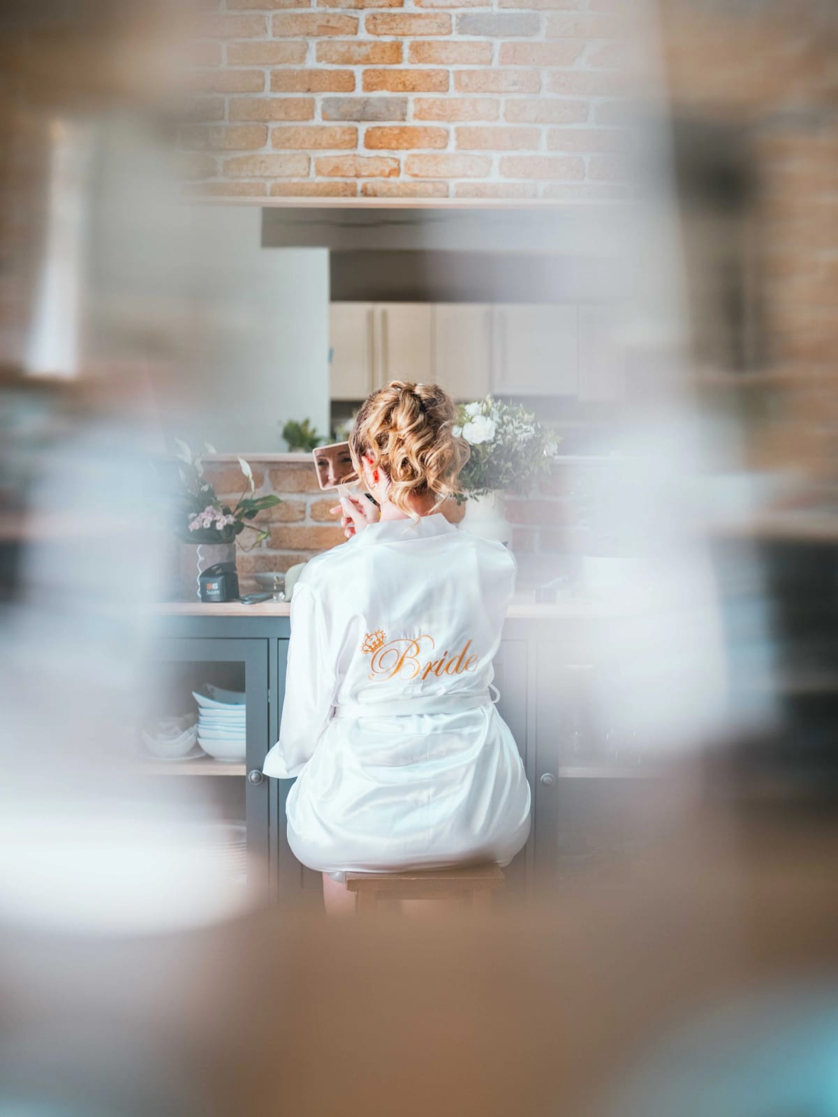 Lydia de dos en peignoir Bride devant le miroir, cheveux bouclés fleuris, pendant ses préparatifs, mariage au Domaine la Ferme du Roy, par Jean-Baptiste Argyre (Jaysphotography)