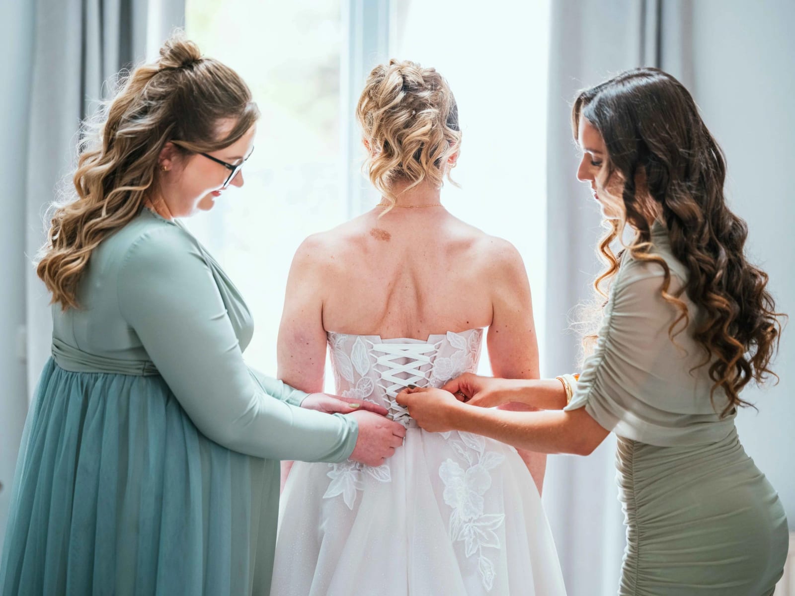 Deux témoins lacent le corset de la robe de Lydia à contre-jour devant la fenêtre, mariage au Domaine la Ferme du Roy, par Jean-Baptiste Argyre (Jaysphotography)