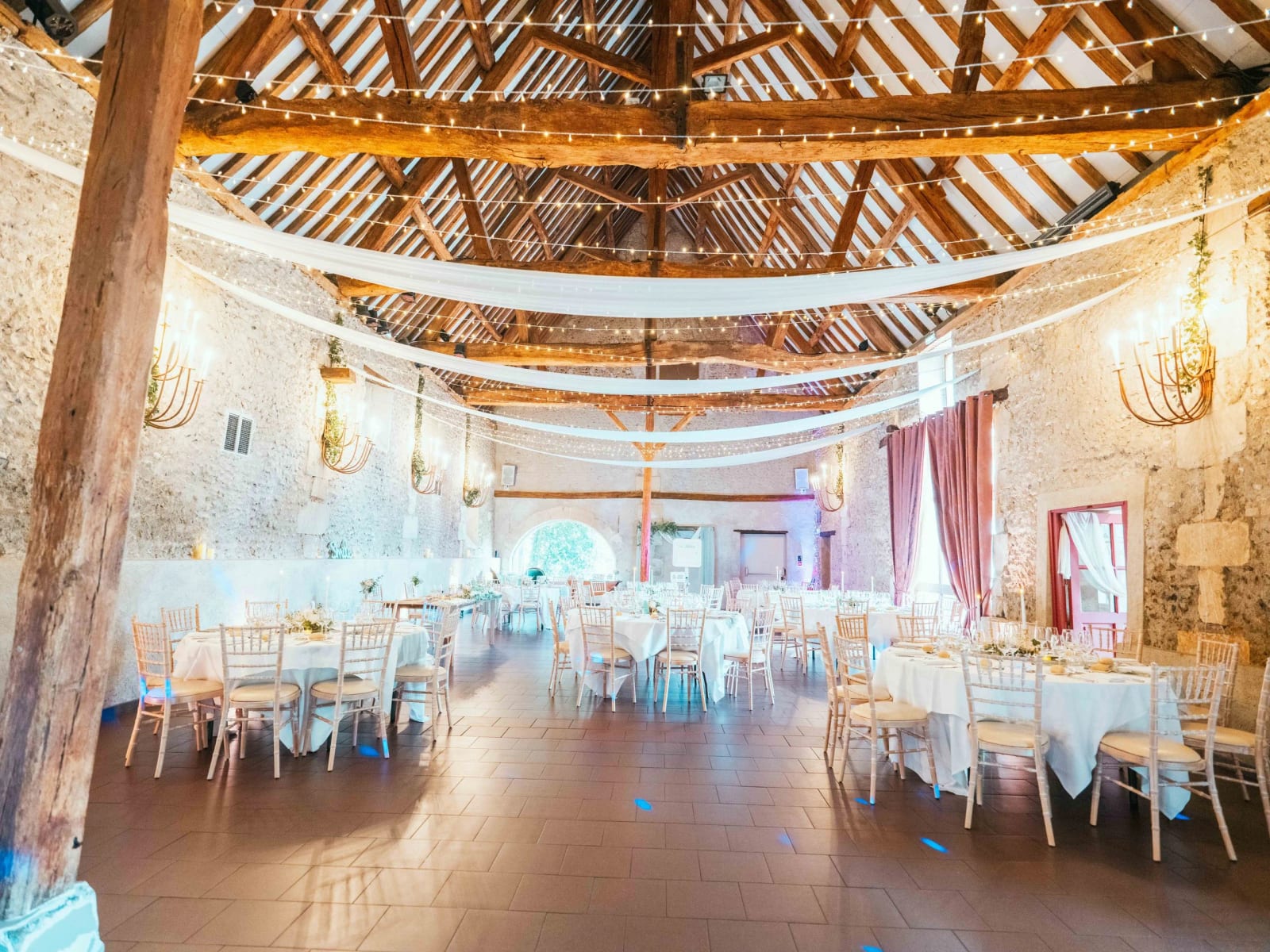 La grange en pierre du Domaine la Ferme du Roy, charpente apparente, guirlandes et tables dressées, mariage au Domaine la Ferme du Roy, par Jean-Baptiste Argyre (Jaysphotography)