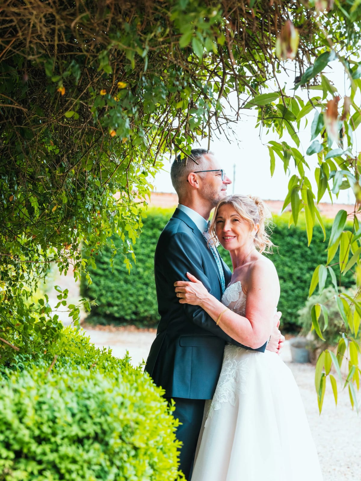 Lydia et Éric enlacés sous un rideau de branches dans le jardin arboré du domaine, mariage au Domaine la Ferme du Roy, par Jean-Baptiste Argyre (Jaysphotography)