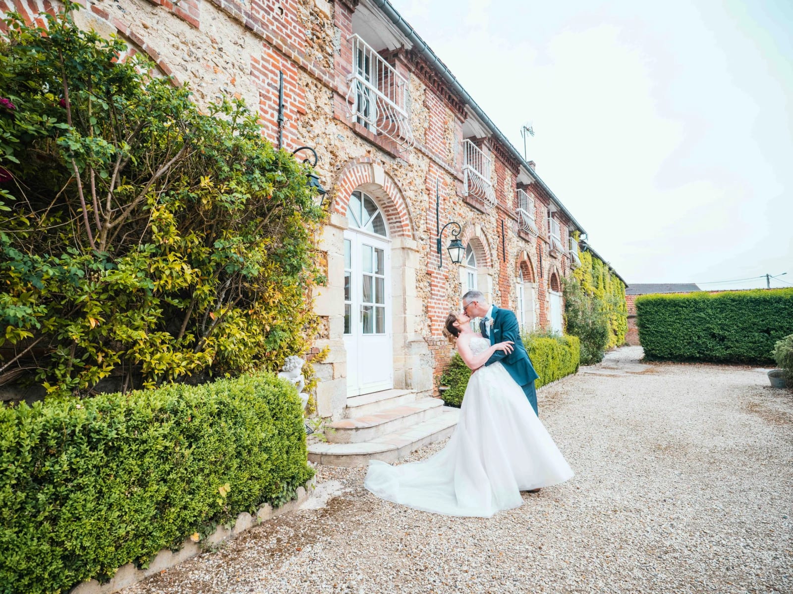 Éric soulève Lydia dans un baiser devant la façade en briques du Domaine la Ferme du Roy, mariage au Domaine la Ferme du Roy, par Jean-Baptiste Argyre (Jaysphotography)