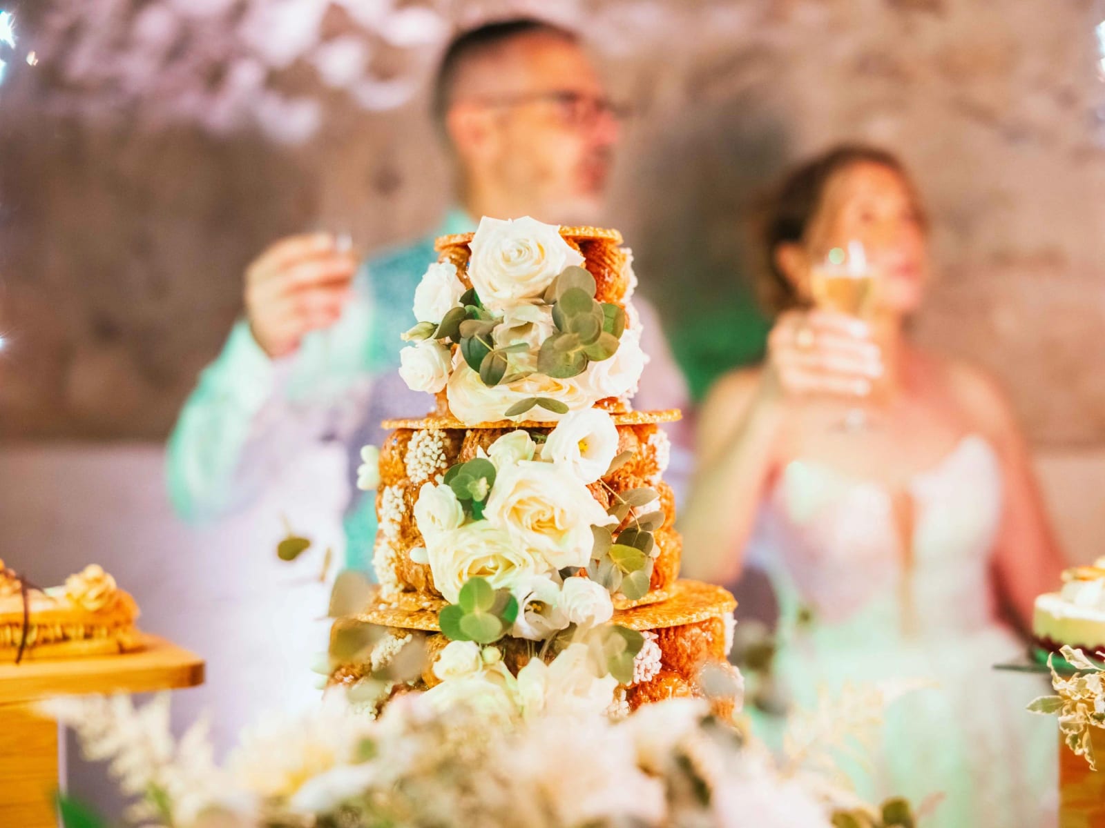 La pièce montée en croquembouche fleurie de roses blanches, les mariés en arrière-plan, mariage au Domaine la Ferme du Roy, par Jean-Baptiste Argyre (Jaysphotography)