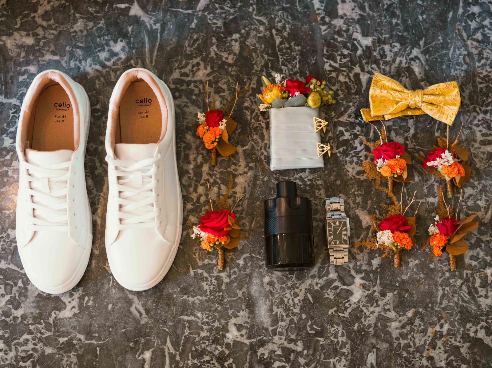 Accessoires du marié sur un marbre gris : baskets blanches Celio, nœud papillon moutarde, montre, parfum, boutons de manchette dorés en forme de Reliques de la Mort et boutonnières de roses rouges, mariage à l'Abbaye de Longpont, par Jean-Baptiste Argyre (Jaysphotography)