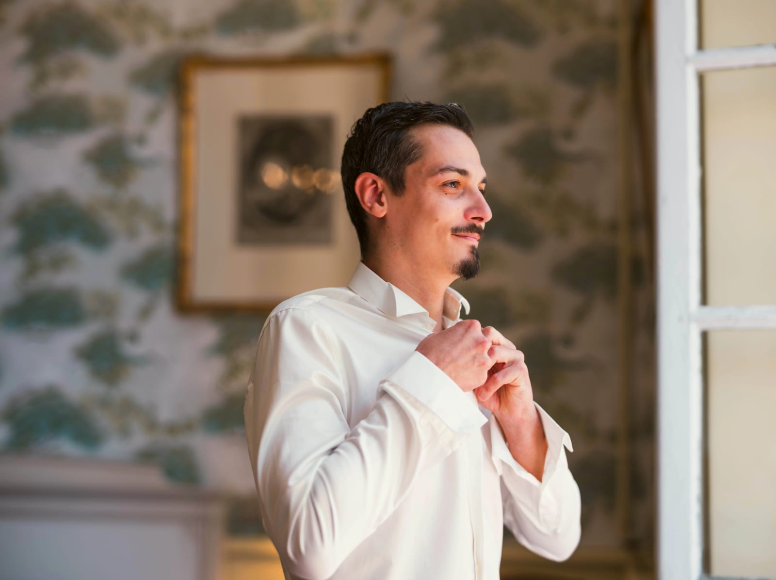 Le marié ferme sa chemise blanche devant un papier peint vert à motifs d'arbres, Abbaye de Longpont, mariage à l'Abbaye de Longpont, par Jean-Baptiste Argyre (Jaysphotography)