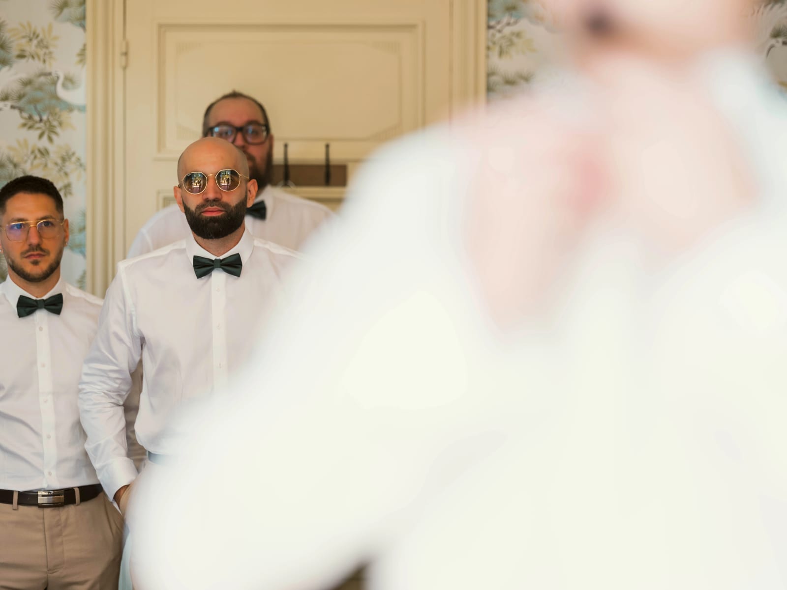 Les amis du marié en chemise et nœud papillon le regardent depuis l'encadrement de la porte, le marié flou au premier plan, mariage à l'Abbaye de Longpont, par Jean-Baptiste Argyre (Jaysphotography)