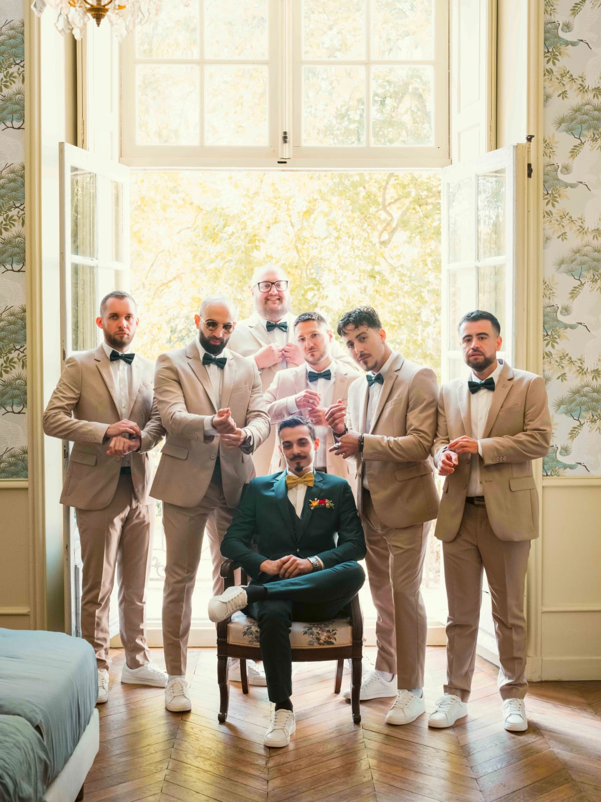 Le marié en costume vert sapin assis devant une porte-fenêtre ouverte, entouré de sept garçons d'honneur en costumes beige, mariage à l'Abbaye de Longpont, par Jean-Baptiste Argyre (Jaysphotography)
