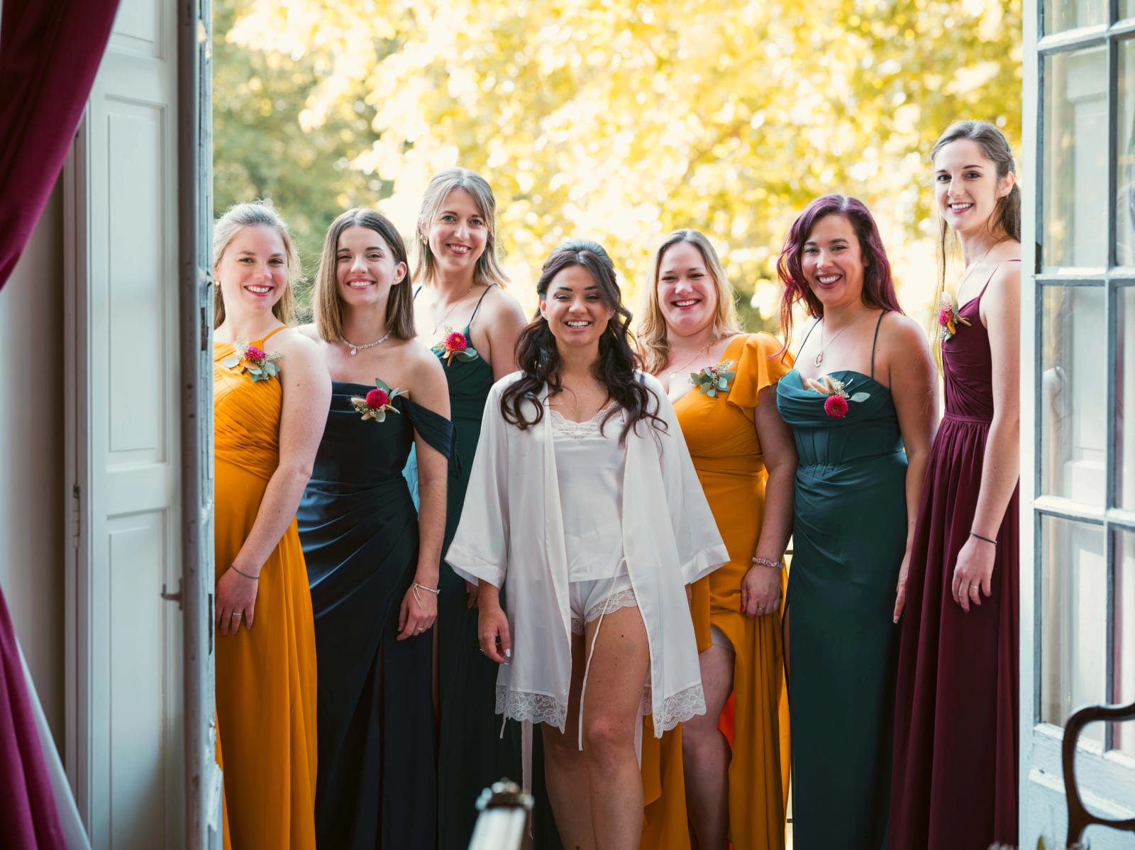 La mariée en kimono blanc entourée de ses demoiselles d'honneur en robes moutarde, vert et bordeaux devant une fenêtre ouverte, mariage à l'Abbaye de Longpont, par Jean-Baptiste Argyre (Jaysphotography)