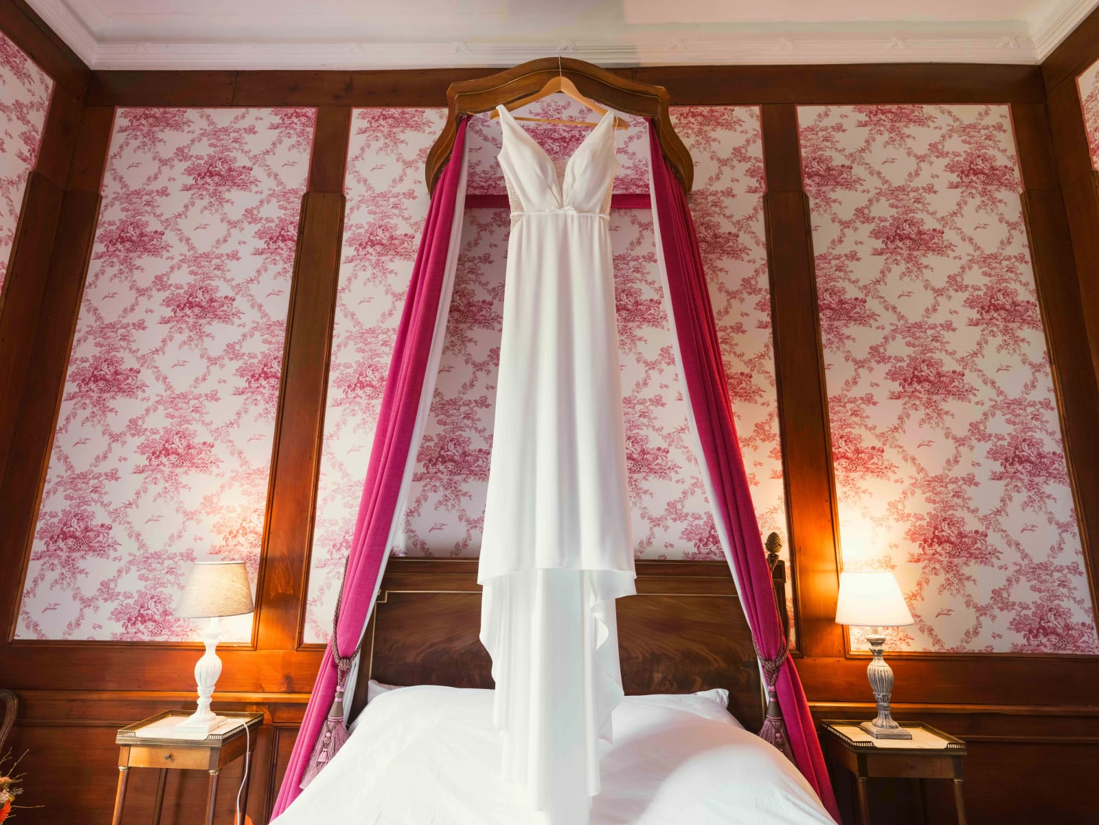 La robe de mariée suspendue au baldaquin d'un lit aux tentures fuchsia, murs en toile de Jouy rose, mariage à l'Abbaye de Longpont, par Jean-Baptiste Argyre (Jaysphotography)