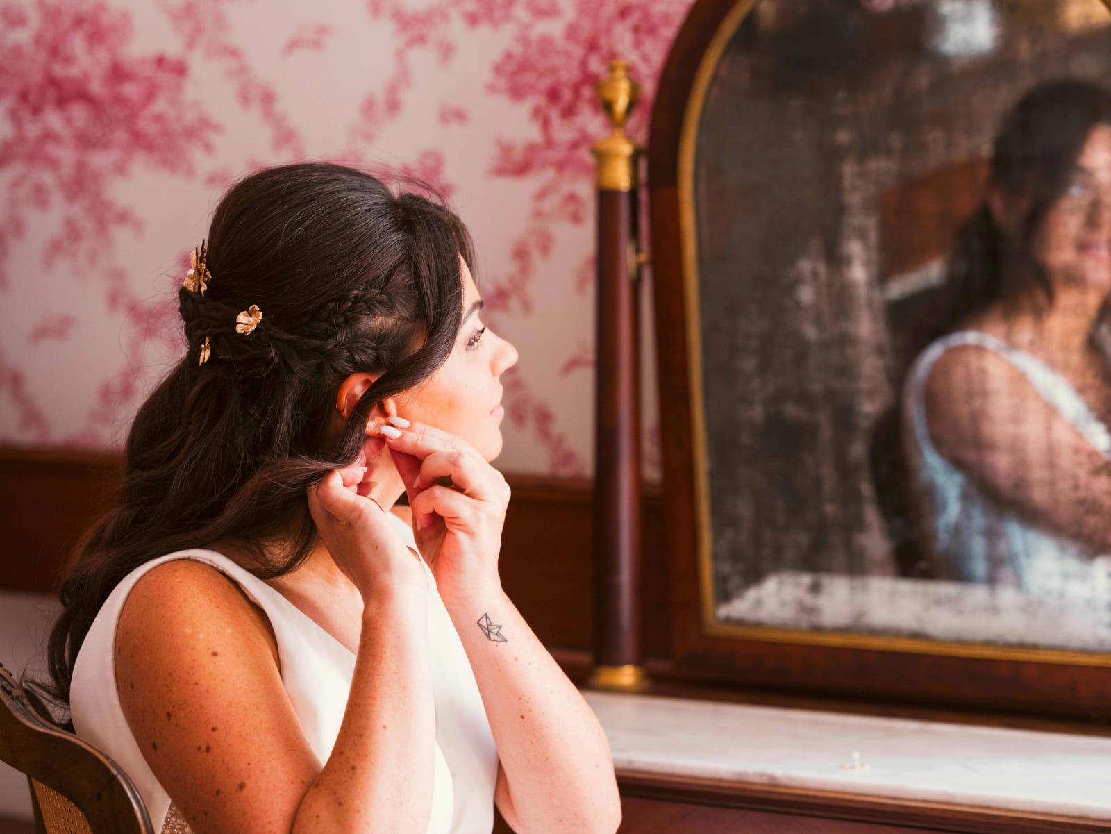 La mariée met ses boucles d'oreilles devant un vieux miroir, cheveux tressés ornés de fleurs dorées, mariage à l'Abbaye de Longpont, par Jean-Baptiste Argyre (Jaysphotography)