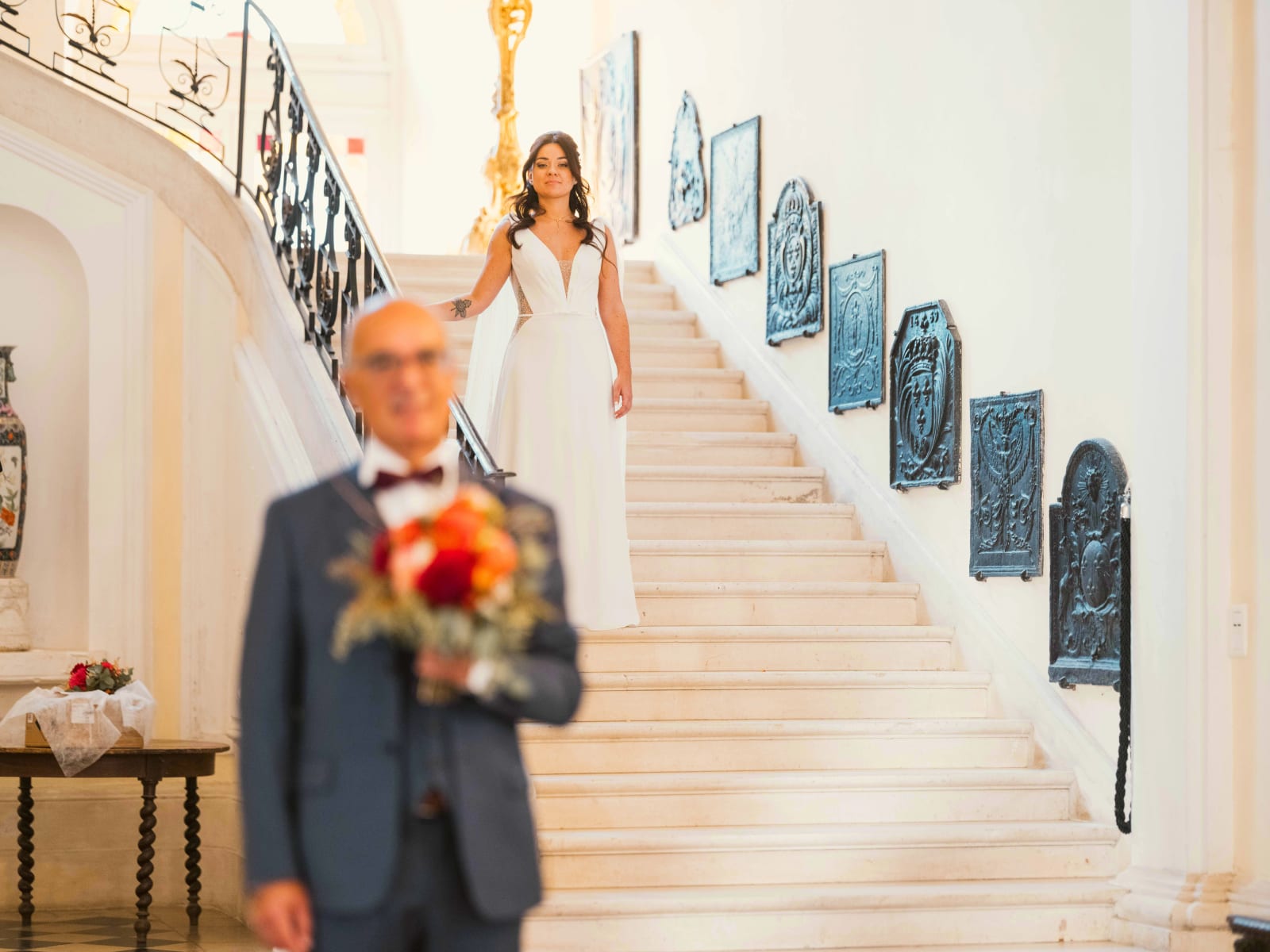 La mariée en haut du grand escalier, un homme en costume gris l'attend en bas avec son bouquet, plaques de cheminée en fonte au mur, mariage à l'Abbaye de Longpont, par Jean-Baptiste Argyre (Jaysphotography)