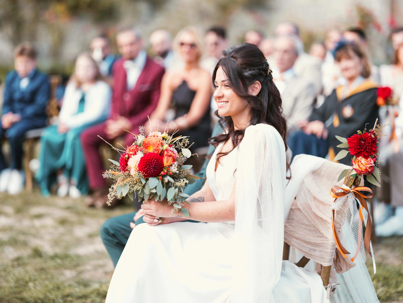 La mariée assise pendant la cérémonie laïque, bouquet de dahlias sur les genoux, invités en arrière-plan, mariage à l'Abbaye de Longpont, par Jean-Baptiste Argyre (Jaysphotography)