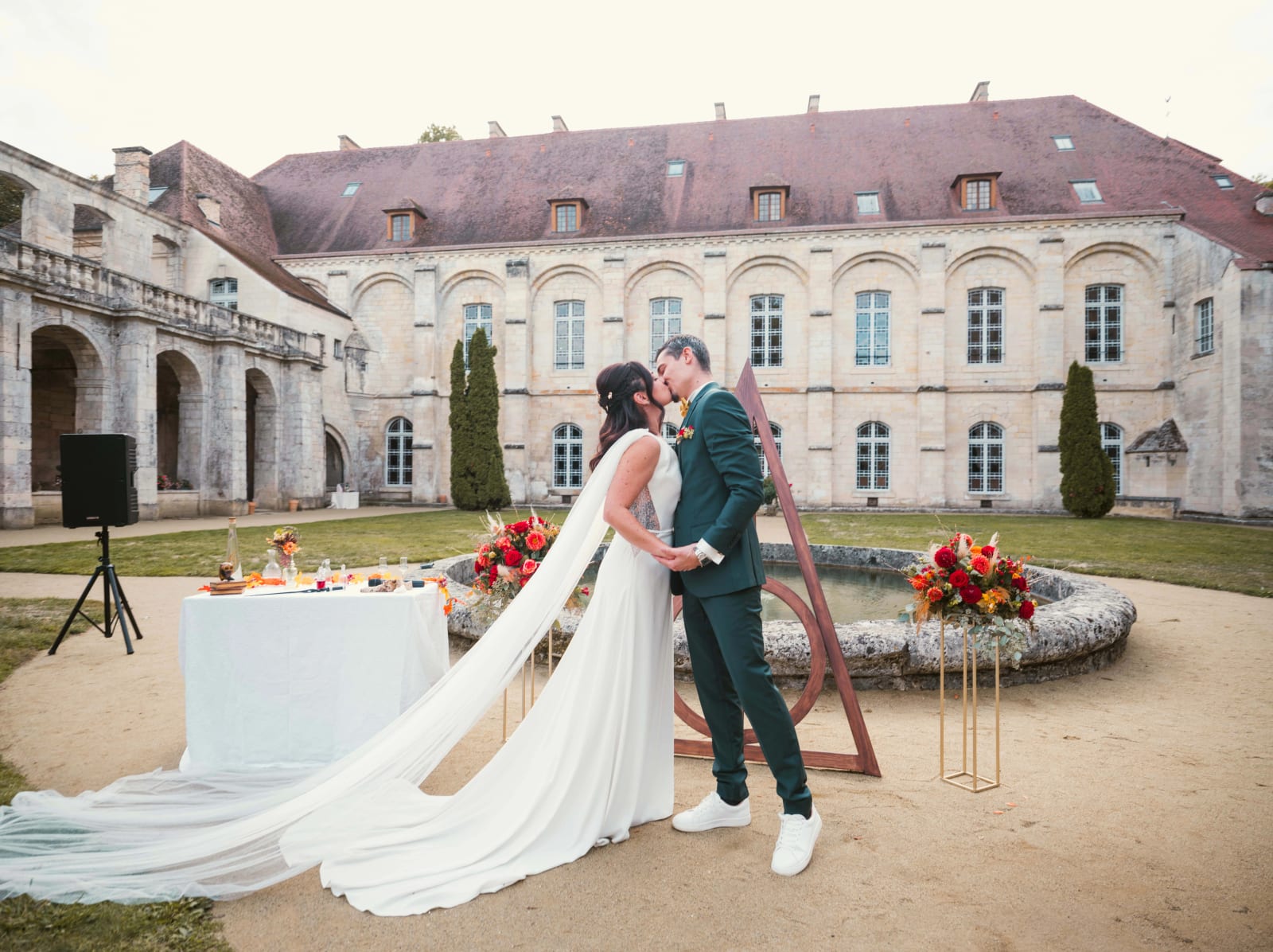 Baiser des mariés devant une arche de bois triangulaire et le bassin de la cour de l'Abbaye de Longpont, longue traîne du voile, mariage à l'Abbaye de Longpont, par Jean-Baptiste Argyre (Jaysphotography)