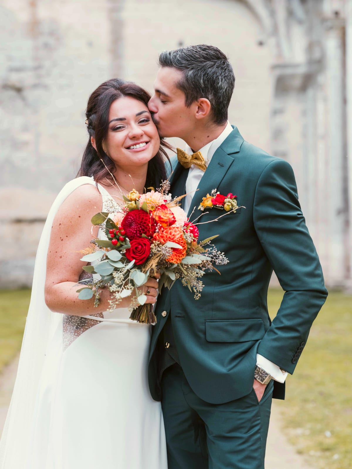Le marié embrasse la mariée sur la joue devant les murs de pierre des ruines, bouquet de dahlias rouges, mariage à l'Abbaye de Longpont, par Jean-Baptiste Argyre (Jaysphotography)