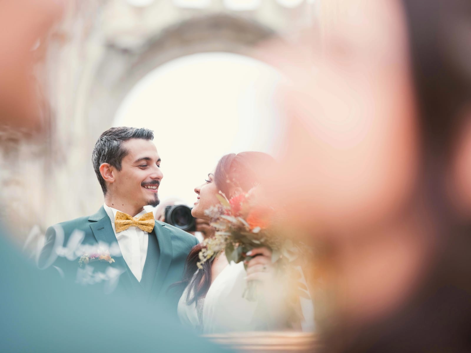 Les mariés entourés d'invités, photographiés entre deux épaules floues, arche des ruines derrière eux, mariage à l'Abbaye de Longpont, par Jean-Baptiste Argyre (Jaysphotography)