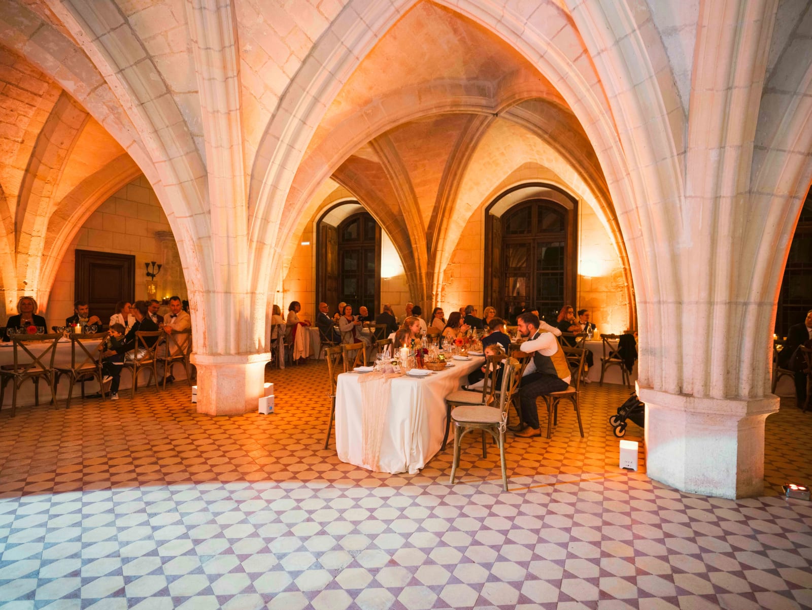 La salle de réception voûtée d'ogives de l'Abbaye de Longpont pendant le dîner, sol carrelé et lumière orangée, mariage à l'Abbaye de Longpont, par Jean-Baptiste Argyre (Jaysphotography)
