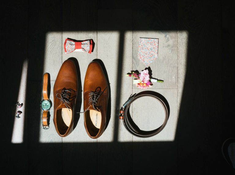 Accessoires de Maxime dans un rai de lumière au Manoir des Brumes : chaussures en cuir, montre, nœud papillon corail, pochette fleurie et ceinture