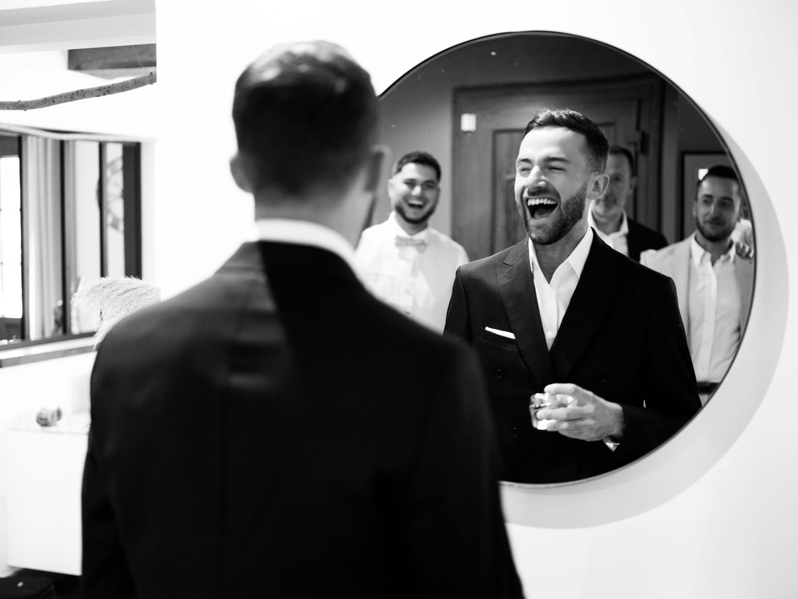 Paul en costume éclate de rire devant un miroir rond, entouré de ses témoins pendant les préparatifs, en noir et blanc, mariage au Château de Moliens, par Jean-Baptiste Argyre (Jaysphotography)