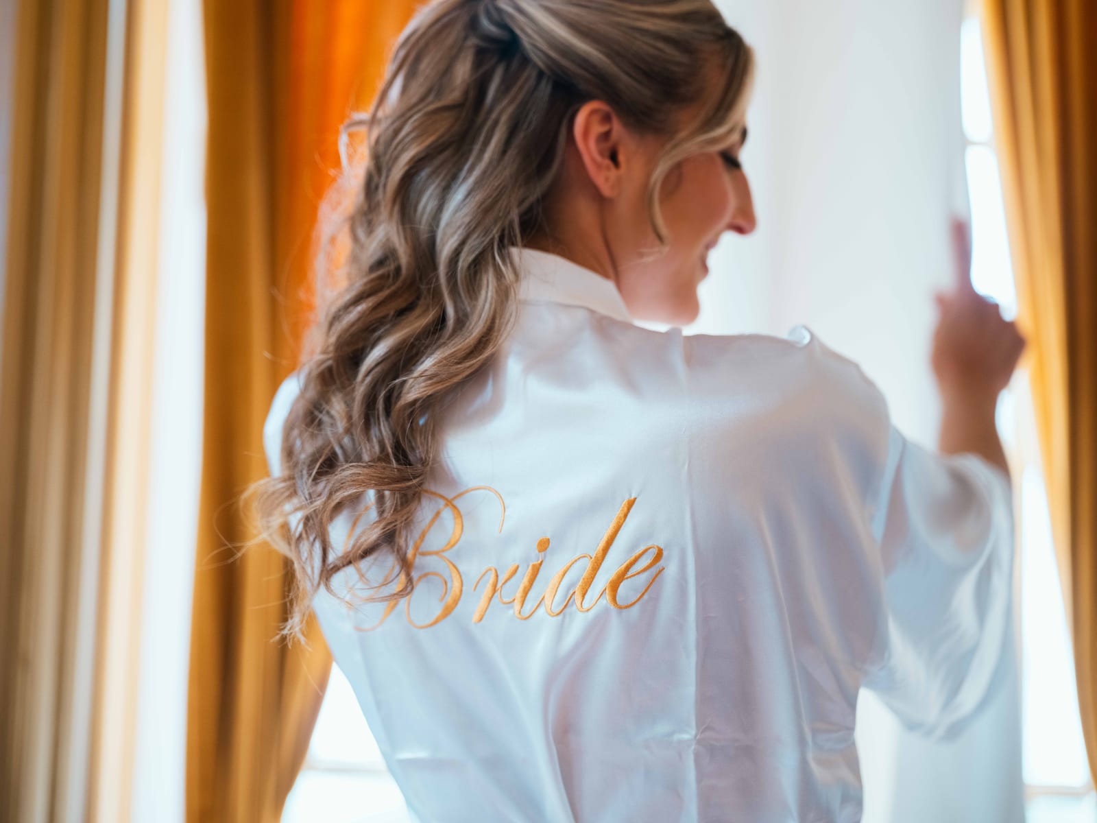 Mathilde de dos en peignoir brodé Bride devant la fenêtre aux rideaux ocre de sa suite au Château de Moliens, mariage au Château de Moliens, par Jean-Baptiste Argyre (Jaysphotography)