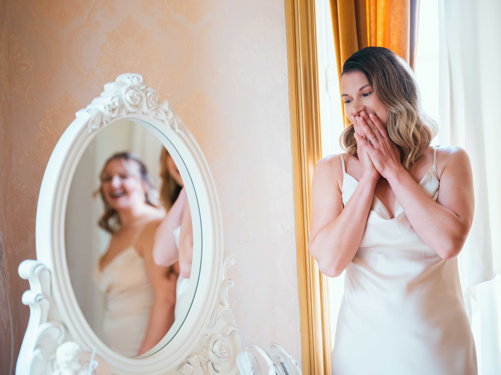 Mathilde émue, les mains sur la bouche, se découvre en robe de mariée dans un miroir baroque au Château de Moliens, mariage au Château de Moliens, par Jean-Baptiste Argyre (Jaysphotography)