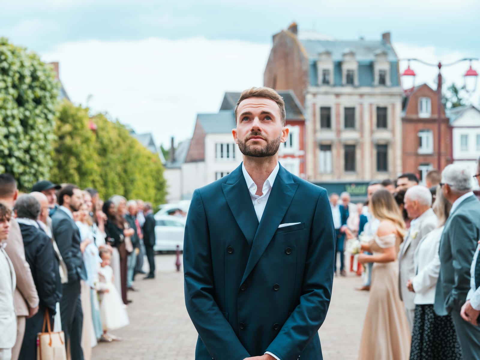 Paul en costume bleu marine attend Mathilde devant la mairie de Crèvecœur-le-Grand, les invités formant une haie sur la place, mariage au Château de Moliens, par Jean-Baptiste Argyre (Jaysphotography)