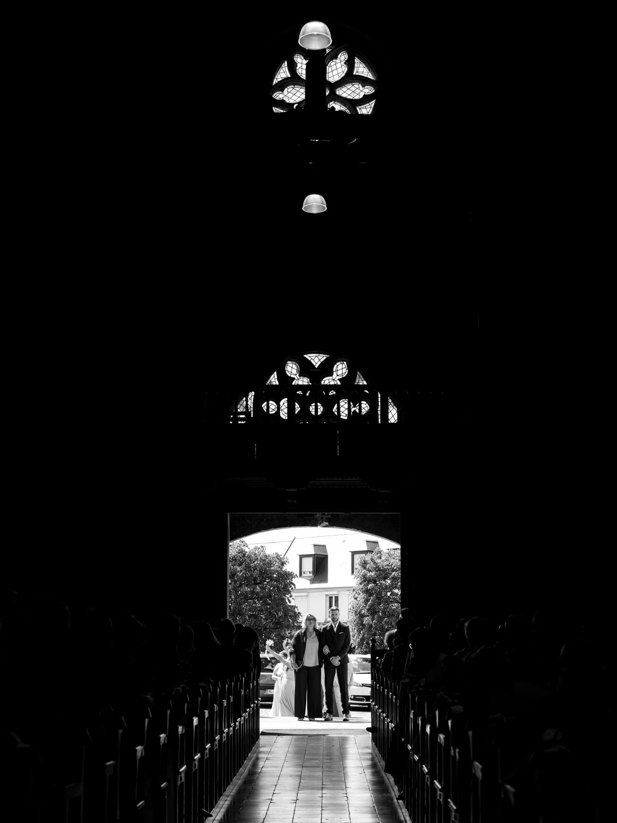 Mathilde au bras de ses proches à l'entrée de l'église de Crèvecœur-le-Grand, silhouettes en contre-jour vues depuis la nef, mariage au Château de Moliens, par Jean-Baptiste Argyre (Jaysphotography)