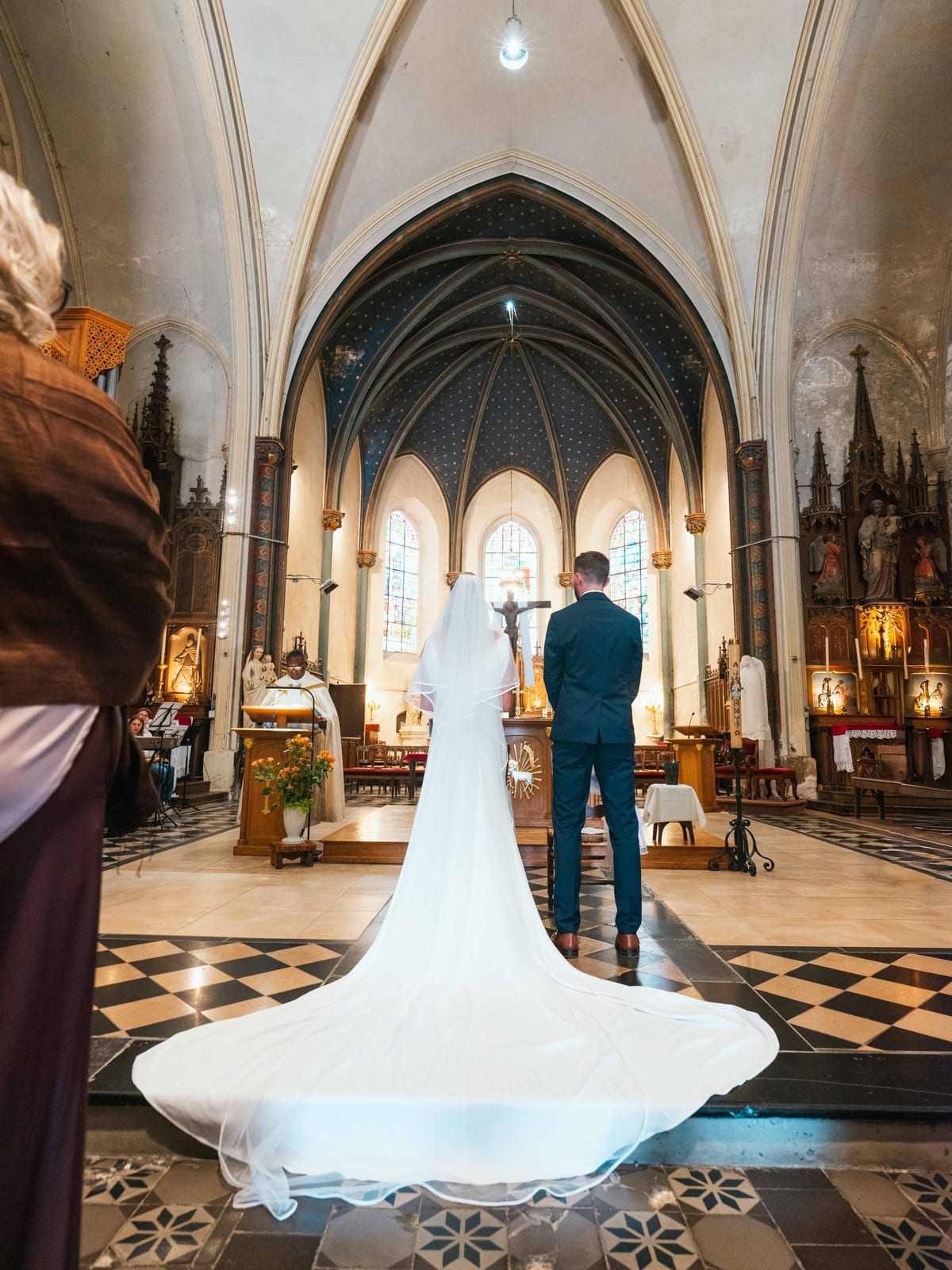 Mathilde et Paul devant l'autel sous les voûtes bleues étoilées de l'église, la traîne de la robe étalée sur le damier, mariage au Château de Moliens, par Jean-Baptiste Argyre (Jaysphotography)
