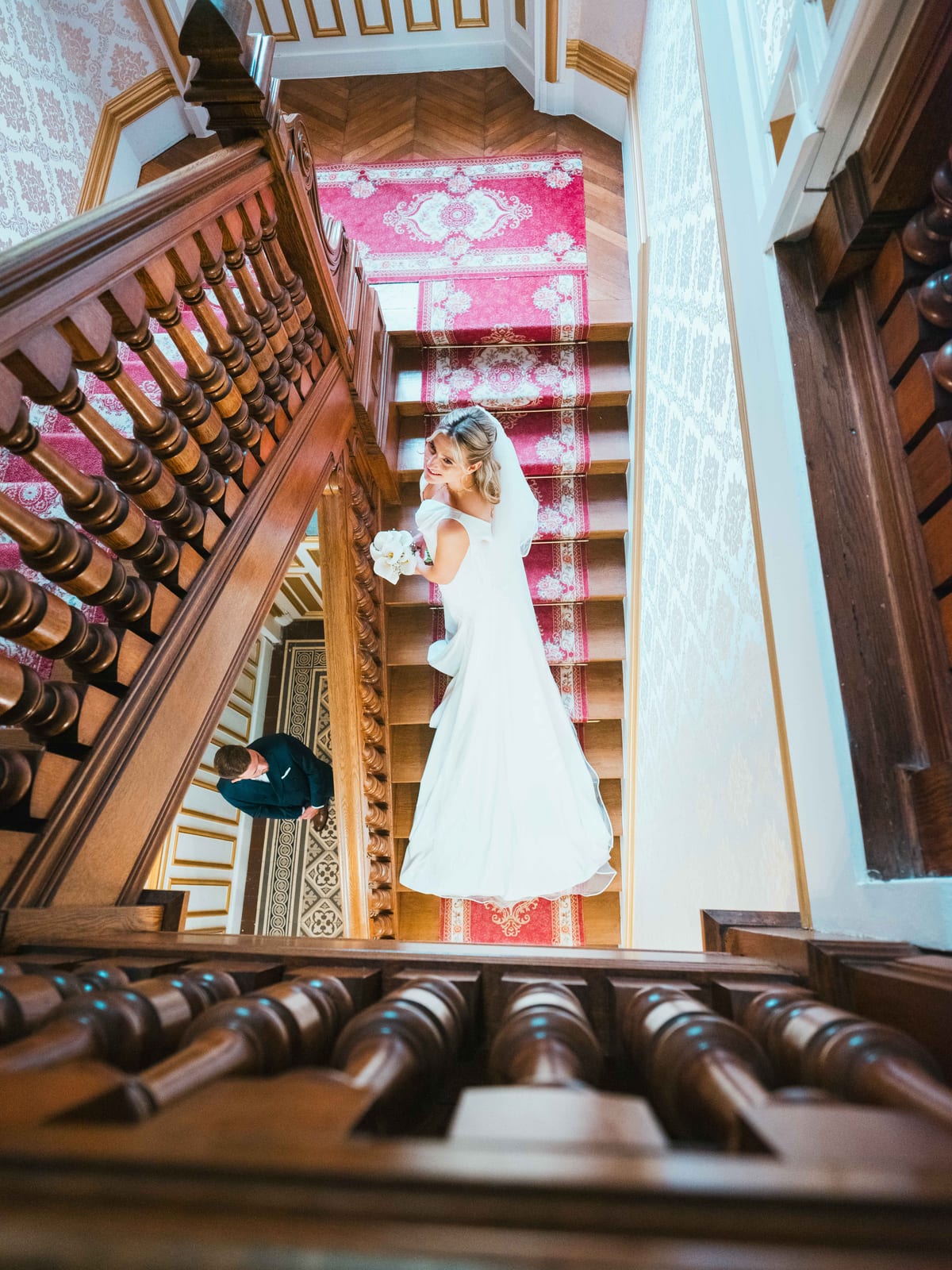 Mathilde en robe de mariée descend le grand escalier en bois du Château de Moliens, bouquet à la main, vue en plongée, mariage au Château de Moliens, par Jean-Baptiste Argyre (Jaysphotography)
