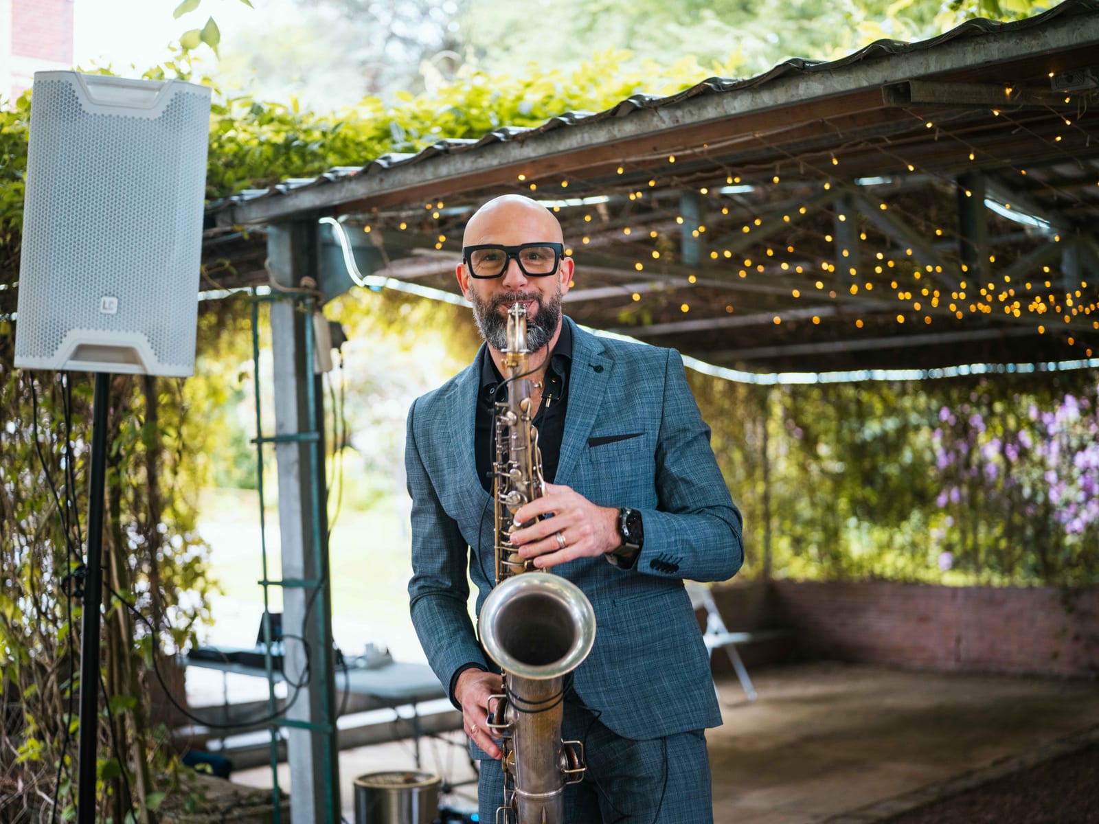 Le saxophoniste joue sous la pergola fleurie et les guirlandes lumineuses pendant le vin d'honneur au Château de Moliens, mariage au Château de Moliens, par Jean-Baptiste Argyre (Jaysphotography)