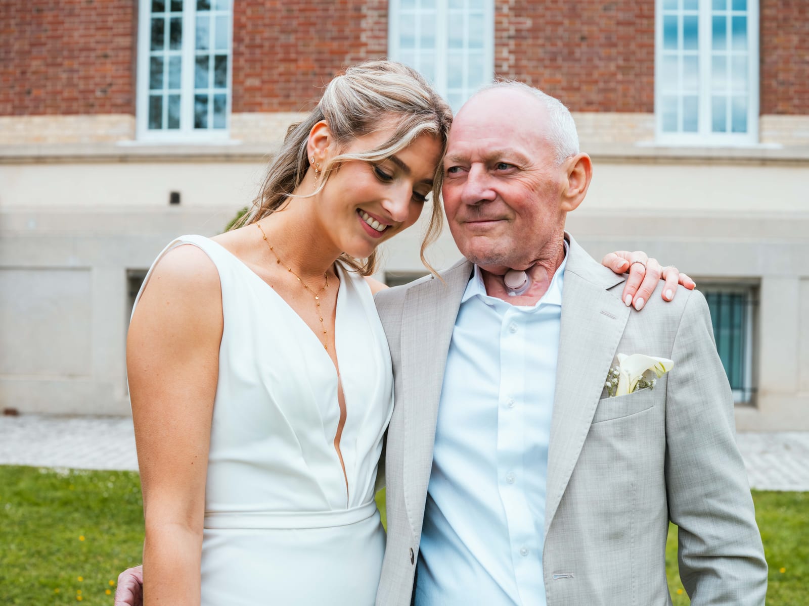 Mathilde joue contre joue avec son papa en costume beige devant la façade en briques du château, mariage au Château de Moliens, par Jean-Baptiste Argyre (Jaysphotography)