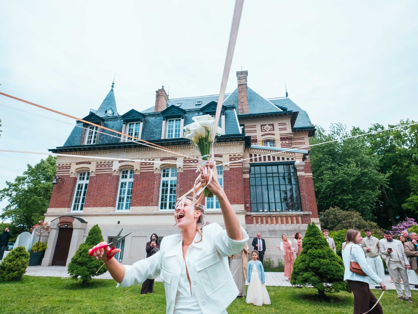 Mathilde lance les rubans colorés pendant la ronde des rubans, les invités en cercle devant le Château de Moliens, mariage au Château de Moliens, par Jean-Baptiste Argyre (Jaysphotography)