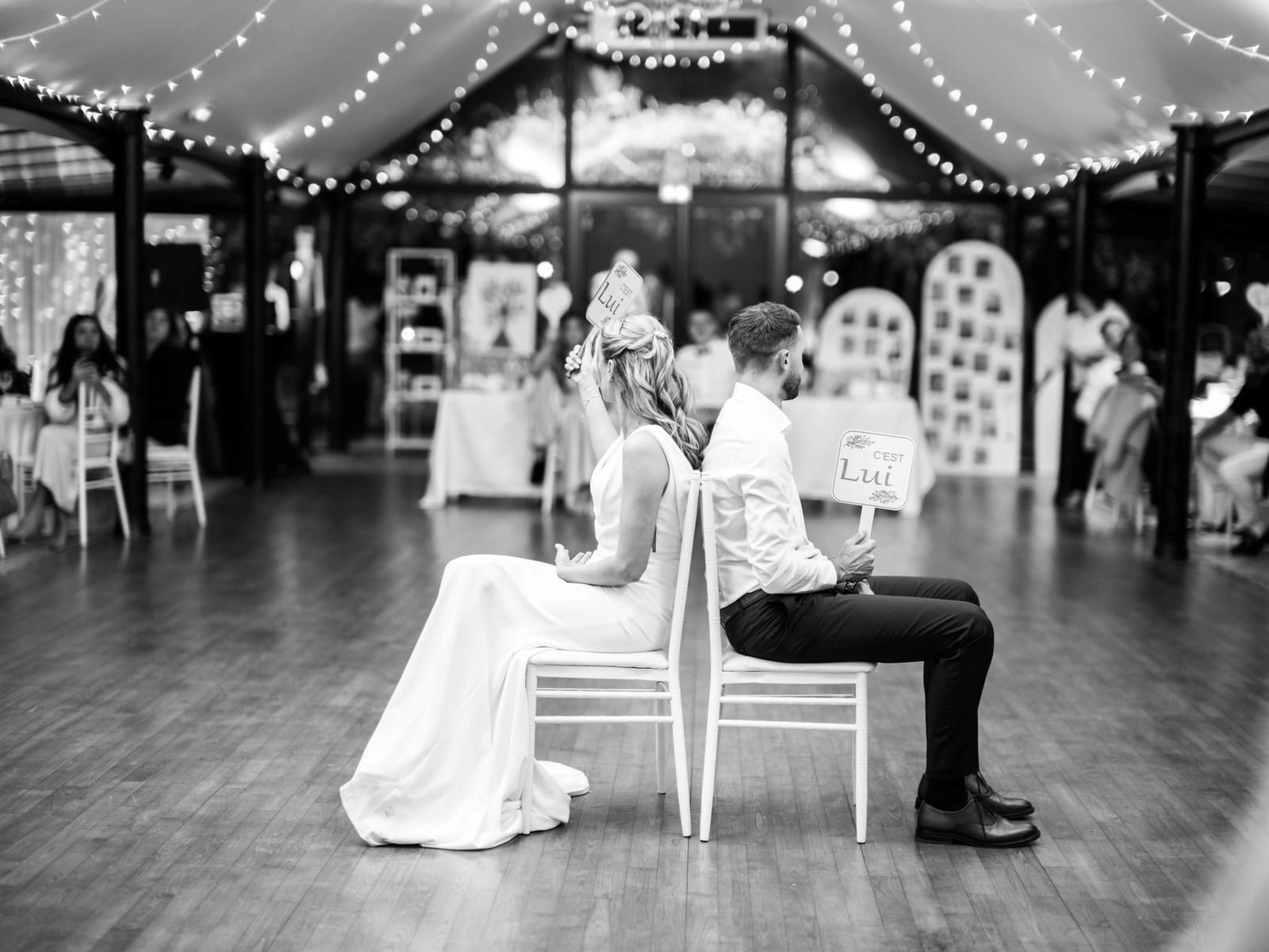 Mathilde et Paul dos à dos pancartes en main pendant le jeu Elle ou Lui de la soirée, en noir et blanc, mariage au Château de Moliens, par Jean-Baptiste Argyre (Jaysphotography)