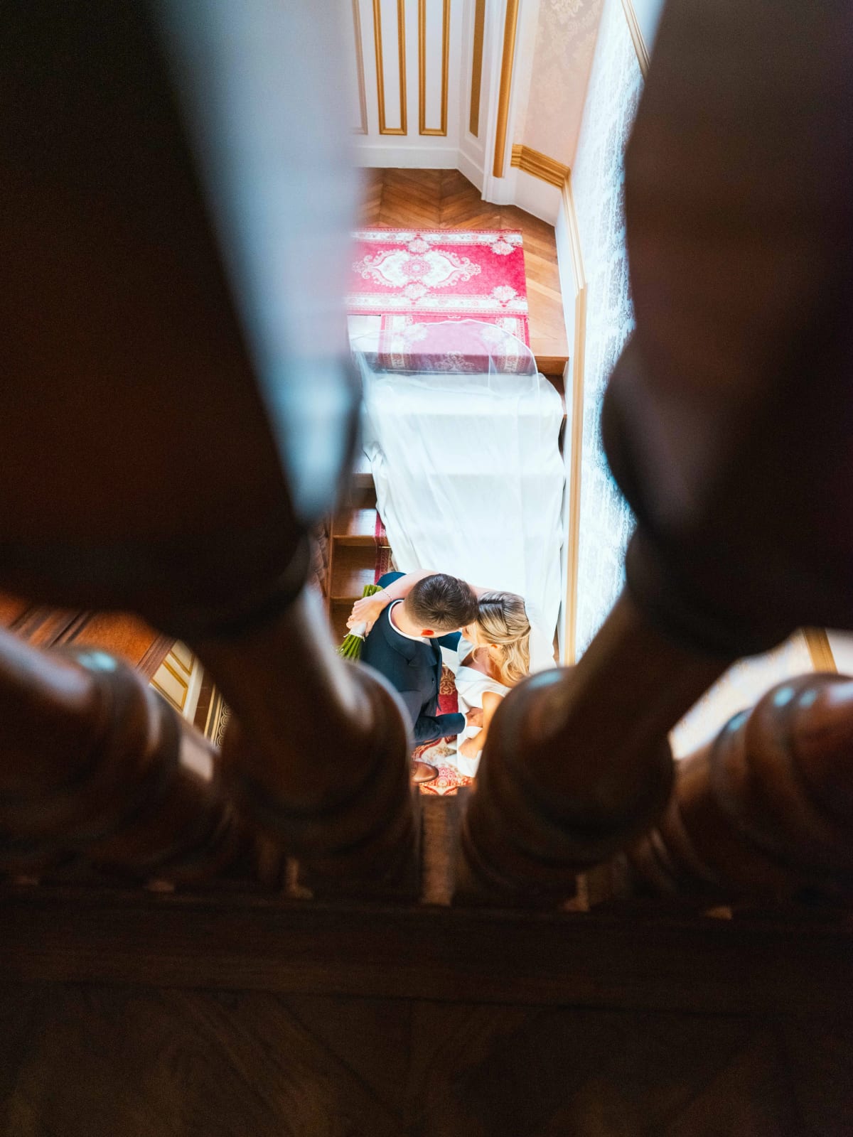 Mathilde et Paul s'embrassent au pied de l'escalier, vus en plongée à travers les barreaux de bois du château, mariage au Château de Moliens, par Jean-Baptiste Argyre (Jaysphotography)