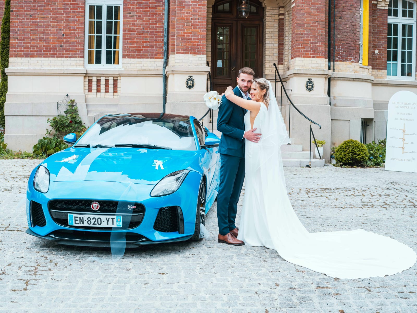 Mathilde et Paul enlacés devant la Jaguar F-Type bleue garée sur les pavés devant la façade du Château de Moliens, mariage au Château de Moliens, par Jean-Baptiste Argyre (Jaysphotography)