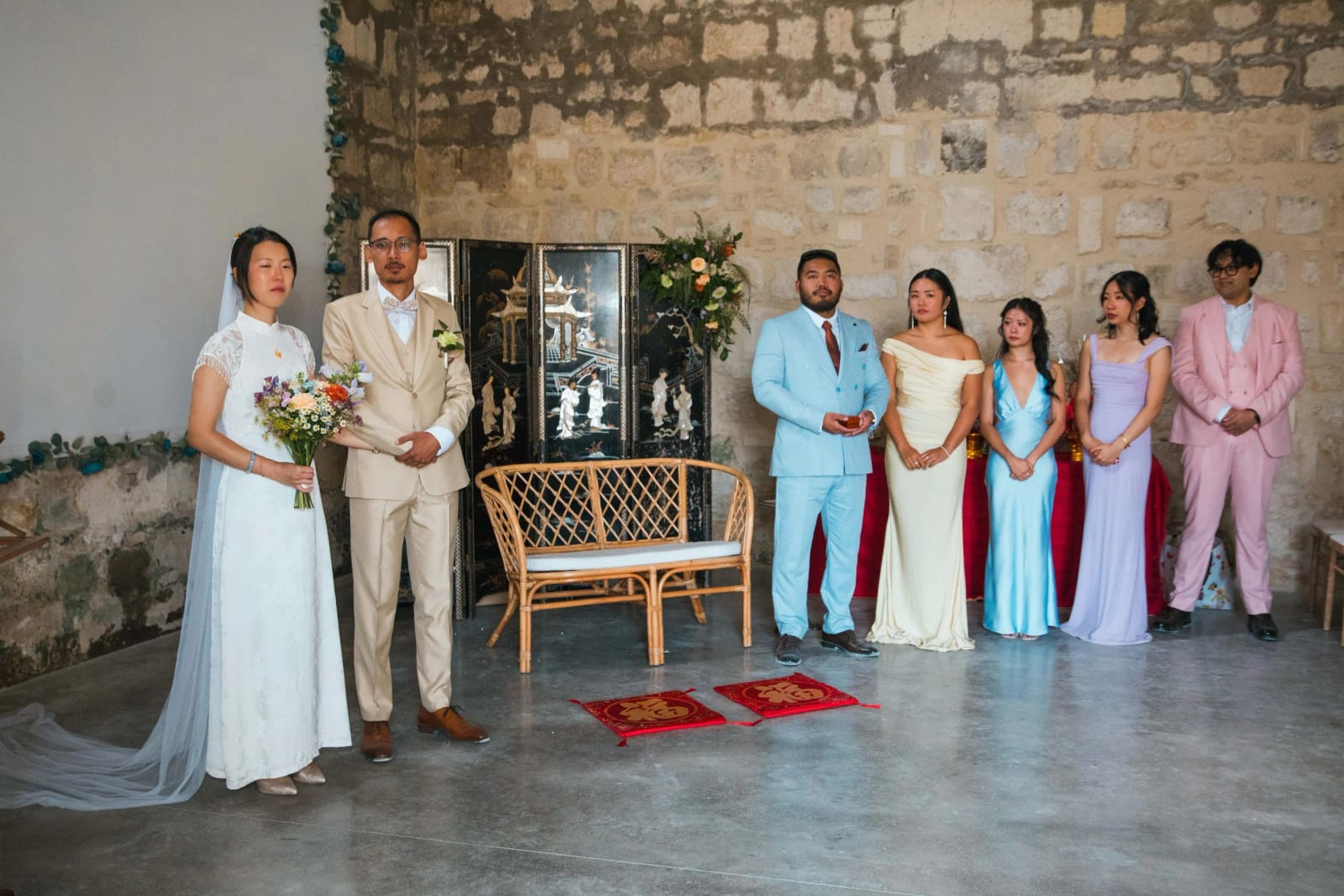 Les mariés debout devant un paravent laqué noir et une banquette en rotin, deux coussins rouges au sol, leurs proches en tenues pastel à côté, mariage au Domaine de Brion, par Jean-Baptiste Argyre (Jaysphotography)