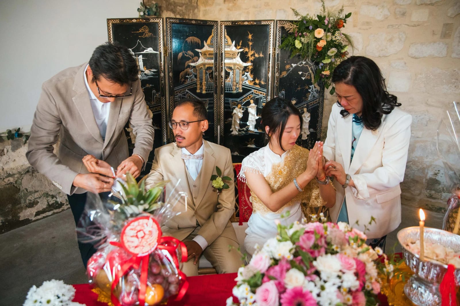 Cérémonie cambodgienne : une proche noue un fil rouge au poignet de la mariée aux mains jointes, le marié assis à côté, paravent laqué derrière, mariage au Domaine de Brion, par Jean-Baptiste Argyre (Jaysphotography)