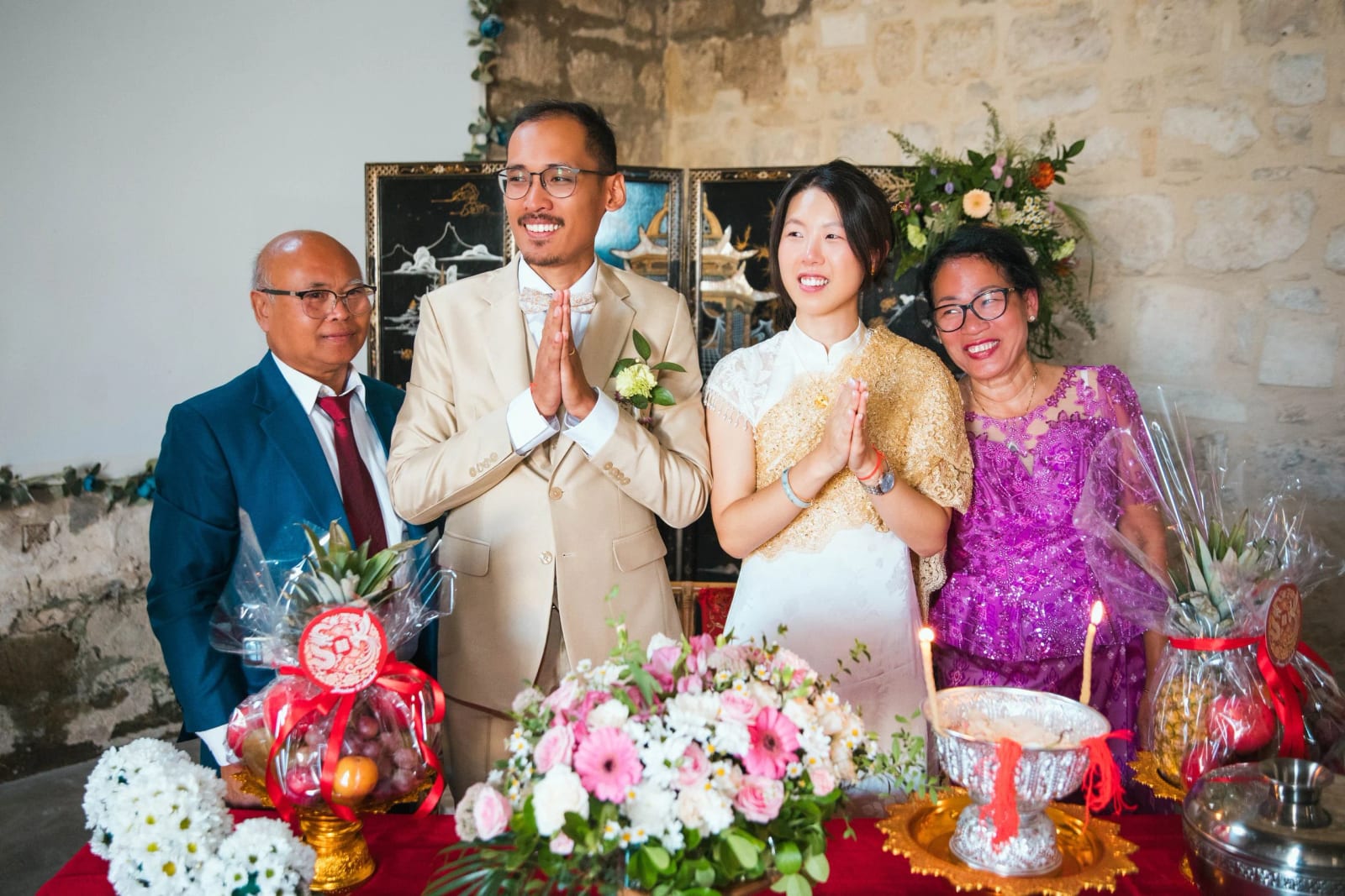 Les mariés mains jointes entre deux proches derrière une table nappée de rouge, corbeilles de fruits, fleurs roses et coupe d'argent, mariage au Domaine de Brion, par Jean-Baptiste Argyre (Jaysphotography)