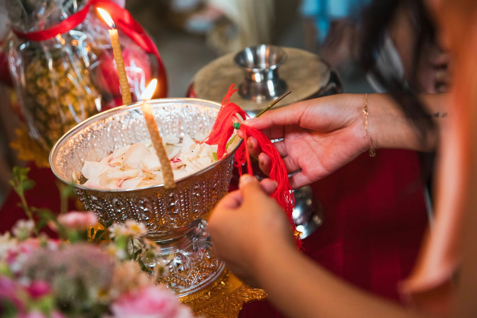 Coupe d'argent ciselé remplie de pétales blancs, deux bougies de cire allumées et des mains qui tiennent des fils rouges, mariage au Domaine de Brion, par Jean-Baptiste Argyre (Jaysphotography)
