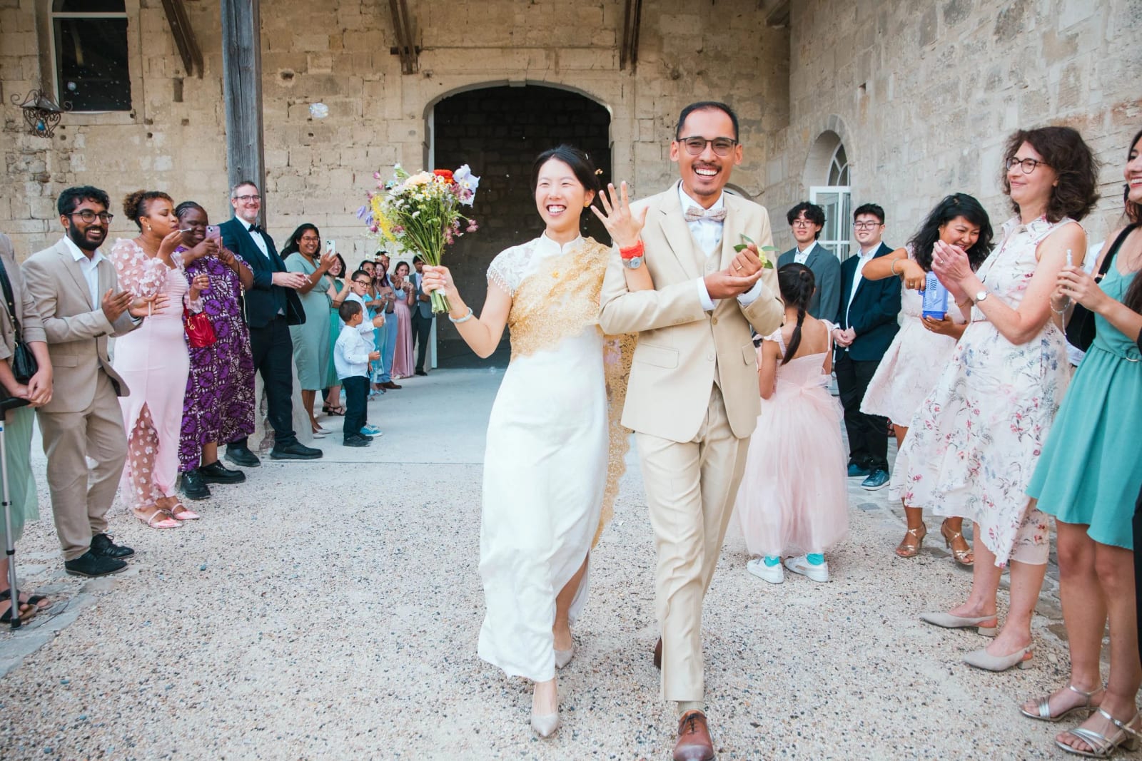 Sortie de cérémonie dans la cour du domaine entre deux haies d'invités qui applaudissent, la mariée lève son bouquet, mariage au Domaine de Brion, par Jean-Baptiste Argyre (Jaysphotography)