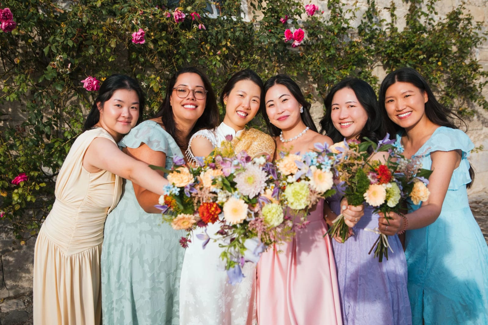 La mariée en étole dorée serrée par ses amies en robes pastel, bouquets de fleurs des champs, devant un mur couvert de rosiers, mariage au Domaine de Brion, par Jean-Baptiste Argyre (Jaysphotography)