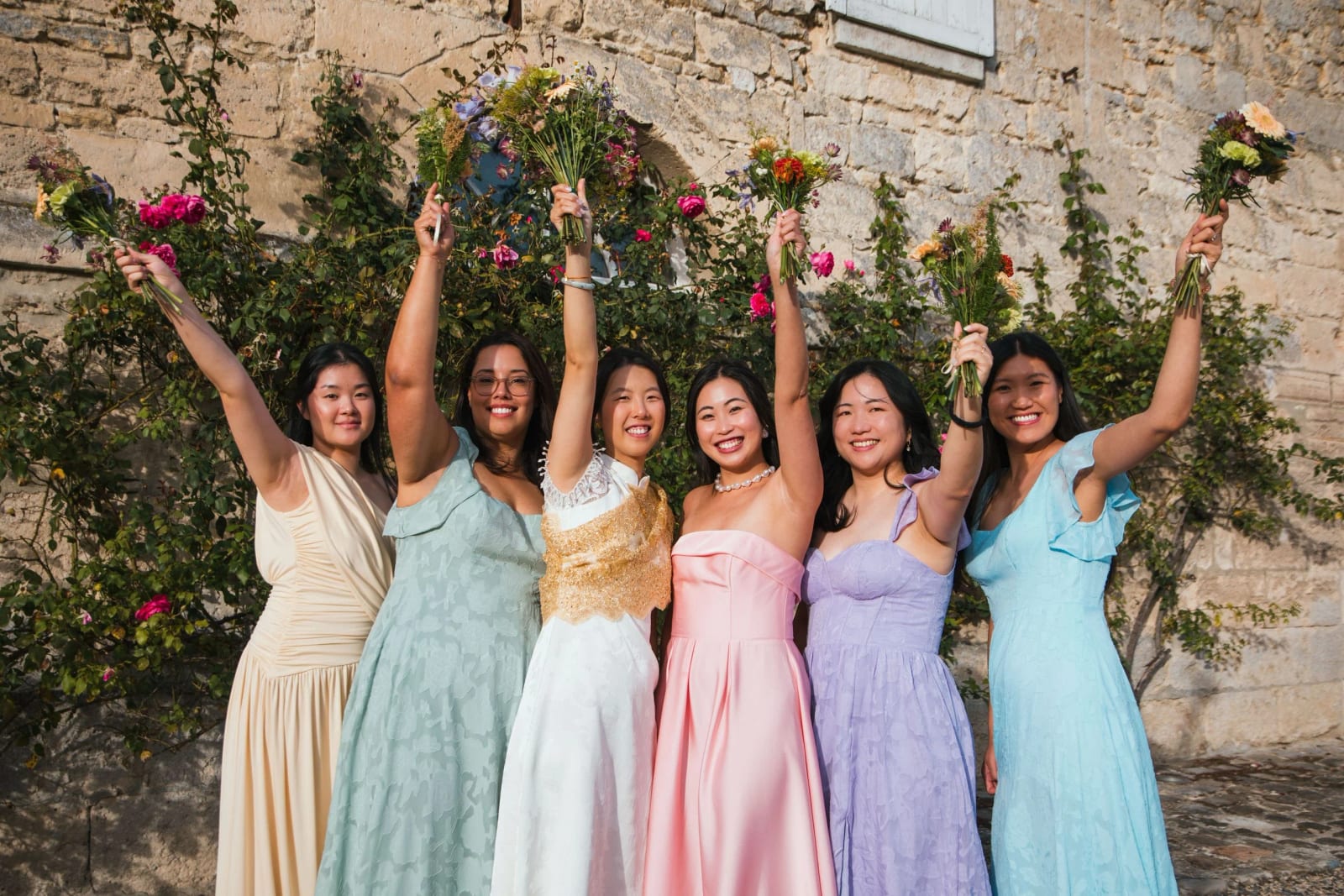 La mariée et ses amies en robes pastel lèvent leurs bouquets devant un mur de pierre et des rosiers, mariage au Domaine de Brion, par Jean-Baptiste Argyre (Jaysphotography)