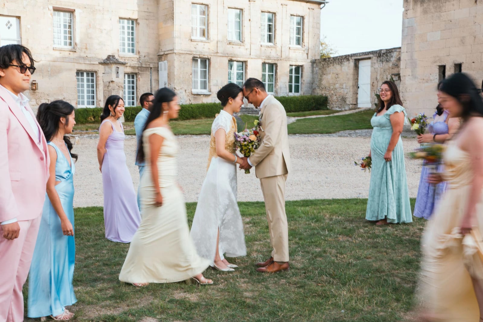 Les mariés front contre front sur la pelouse devant la façade du Domaine de Brion, leurs proches en robes pastel passent autour d'eux en mouvement, mariage au Domaine de Brion, par Jean-Baptiste Argyre (Jaysphotography)