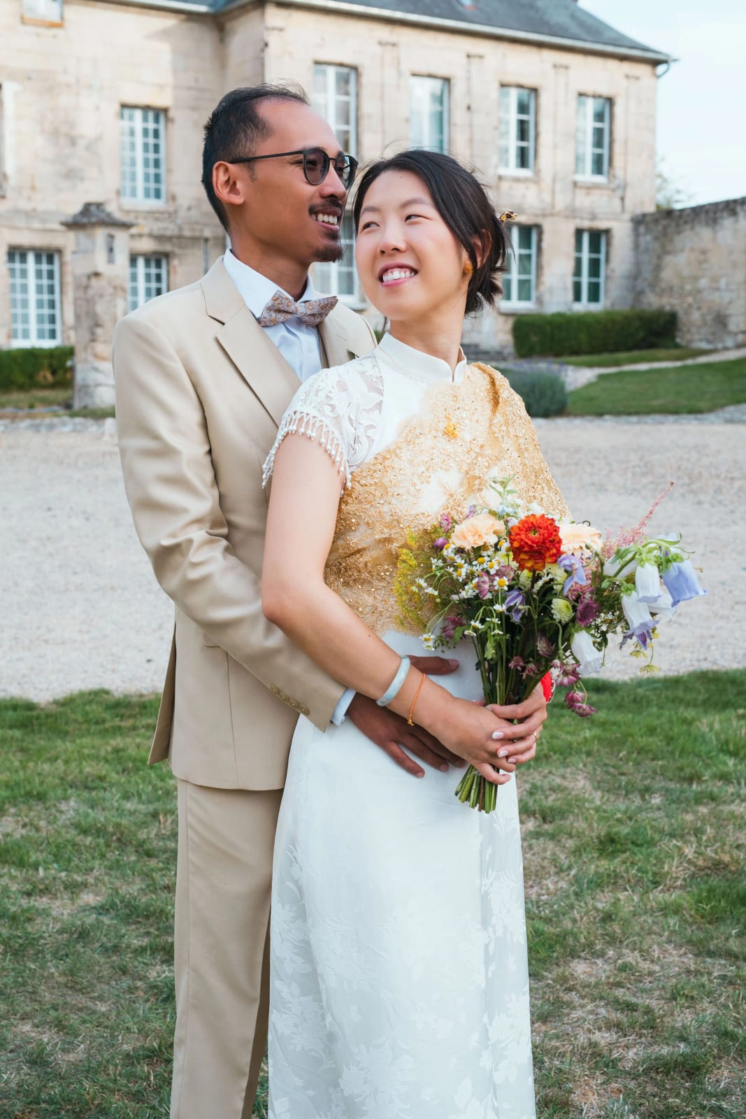 Le marié enlace la mariée par derrière devant la façade aux fenêtres blanches, bouquet de dahlias et fleurs des champs, mariage au Domaine de Brion, par Jean-Baptiste Argyre (Jaysphotography)